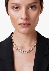 Guldtonet halskæde med firkantede og pyramideformede accenter med klare krystaller, bundet sammen med metal kæde, båret over en sort blazer.