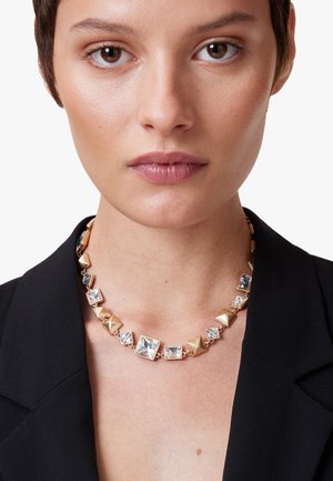 Goudkleurige ketting met vierkante en piramidevormige accenten bezet met heldere kristallen, verbonden met een metalen ketting, gedragen over een zwarte blazer.