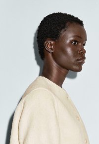 Profilo di una persona con capelli ricci corti che indossa un cappotto di lana color crema su uno sfondo grigio chiaro.