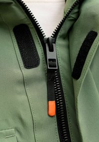 Olivegrüne Jacke mit schwarzem Reißverschluss und orangem Zipper. Verfügt über schwarze Klettverschluss-Patches und eine glatte Stofftextur.