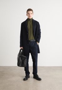 Dunkelblauer Übermantel über einem grünen Rollkragenpullover, kombiniert mit dunklen Hosen und schwarzen Schuhen, während eine schwarze Lederreisetasche gehalten wird.