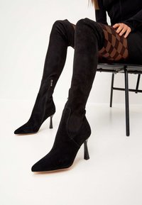 Botas de ante negro hasta la rodilla con punteras afiladas, tacones delgados y cremalleras laterales; superficie texturizada y diseño ajustado.
