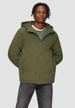 Veste isolée verte avec fermeture éclair et capuche, dotée de poches latérales et d'un ourlet légèrement courbé. Portée par-dessus un pull texturé.