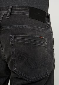 Jeans de mezclilla negra con un parche de cuero que lleva la etiqueta "TOM TAILOR". Patrón de costura visible en los bolsillos traseros.