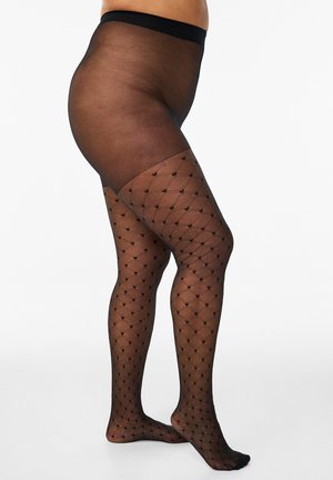 Inimene, kes kannab musta läbipaistvat mustrilisi sukkpükse teemant- ja täpiprindiga, seisab valge tausta ees.