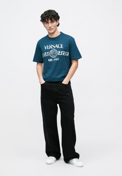 Tirkīzzils kokvilnas T-krekls ar baltu Versace logotipu un lauva grafiku, kopā ar plaša piegriezuma melnām biksēm un baltām sporta kurpēm.