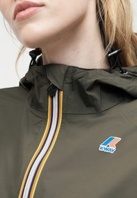 Κλείσιμο πλάνου ενός ατόμου που φοράει ένα χακί μπουφάν με πολύχρωμη ρίγα στο φερμουάρ και το λογότυπο K-WAY στο στήθος.