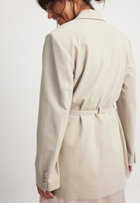 Beige tailored blazer with a belted waist, notched lapels, and three-button cuffs. Smooth texture and structured fit, designed for a polished look.