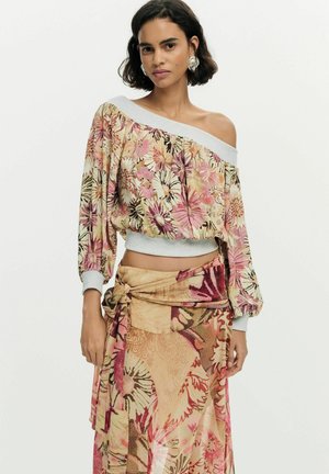 Naine lühikeste tumedate juustega kannab lillelist õlgadeta pluusi ja sobivat mähkseelikut roosa, beeži ja pruuni toonidega valgel taustal.