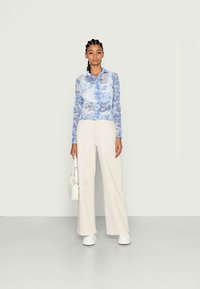 Blusa floral azul transparente com fecho de botão, combinada com calças largas de cintura alta em creme e ténis brancos, segurando uma pequena bolsa branca.