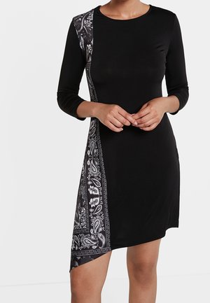 Femme portant une robe noire à manches longues et ourlet asymétrique, avec un motif paisley blanc vertical sur un côté.
