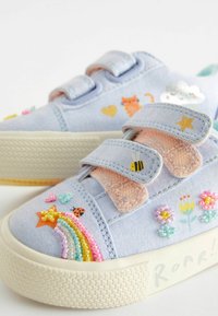 Světle modré plátěné tenisky s suchými zipy, které mají různé barevné vyšívané vzory, včetně duhy, květin a zvířat.