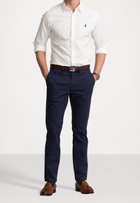 Witte overhemd met knopen, donkerblauwe broek, brown leren riem en brown loafers. Het overhemd heeft een klein logo. Casual fit zonder zichtbare patronen.
