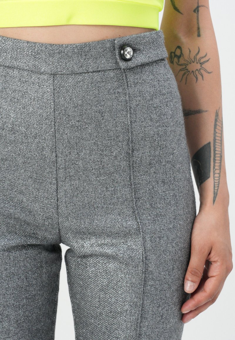Primer plano de pantalones gris de cintura alta con un botón decorativo, combinados con una blusa de color amarillo neón, y un brazo tatuado descansando a un lado.