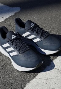Baskets de sport bleu foncé avec des accents blancs et trois rayures, dotées d'un dessus texturé, d'un col rembourré et d'une semelle blanche flexible.