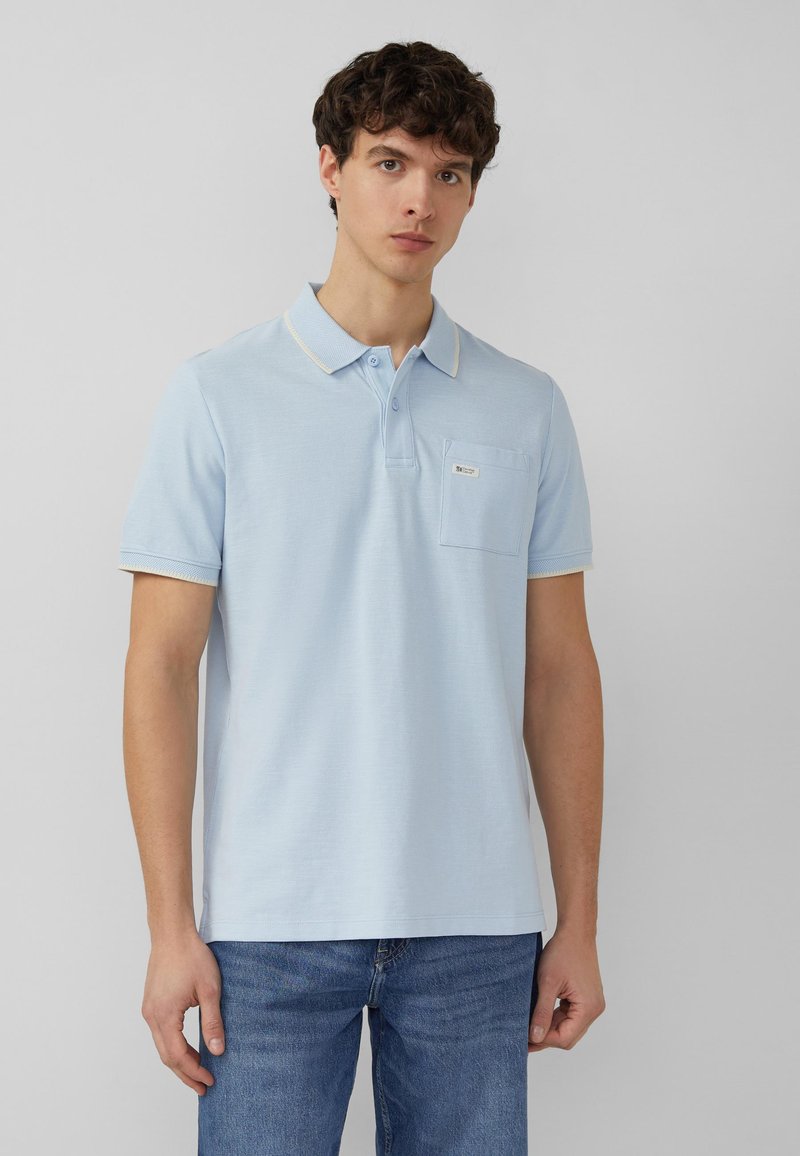 Junger Mann steht, trägt ein hellblaues Polohemd mit kurzen Ärmeln und Brusttasche sowie blaue Jeans vor einem schlichten grauen Hintergrund.