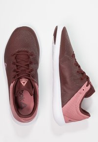 Burgunderfärgade träningsskor med en strukturerad nätöverkonstruktion, vit mellansula och rosa accenter vid hälen och sidan. Har snörningsdesign och logotyp.