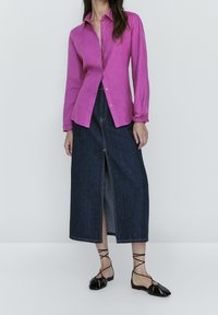Pink long-sleeve button-up shirt paired with a dark denim skirt featuring a front slit and black strappy flat shoes. Smooth fabric textures.
