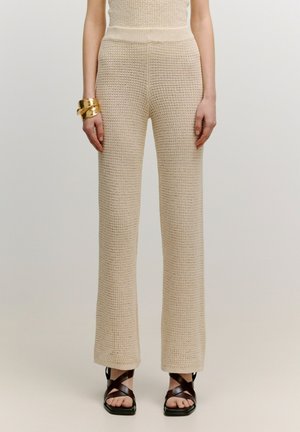 Beige gestrickte weit geschnittene Hosen mit einem strukturierten Muster, hohem Bund und Seitenschlitzen am Saum, kombiniert mit dunkelbraunen Riemchensandalen.