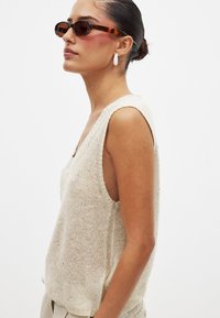Beige knit sleeveless top with a deep V-neck, textured fabric, and an unrefined edge finish. Round tortoiseshell sunglasses and white earrings.