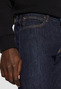 Vaqueros de mezclilla azul oscuro con costuras amarillas, un bolsillo delantero y una textura suave. La cintura está ligeramente elevada y cuenta con herrajes dorados.