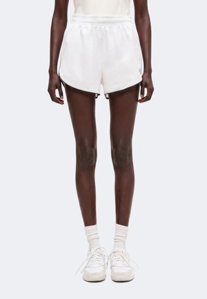Persona in pantaloncini sportivi bianchi, calzini bianchi e scarpe da ginnastica bianche davanti a uno sfondo chiaro e uniforme.