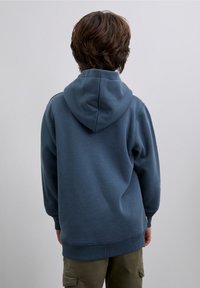 Felpa con cappuccio blu a vestibilità morbida con polsini a costine, mostrata di spalle, abbinata a pantaloni cargo verde oliva.