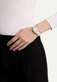 Montre carrée avec un bracelet en silicone blanc, des accents en or rose et un cadran blanc minimaliste. Portée sur une main reposant sur un pantalon noir.