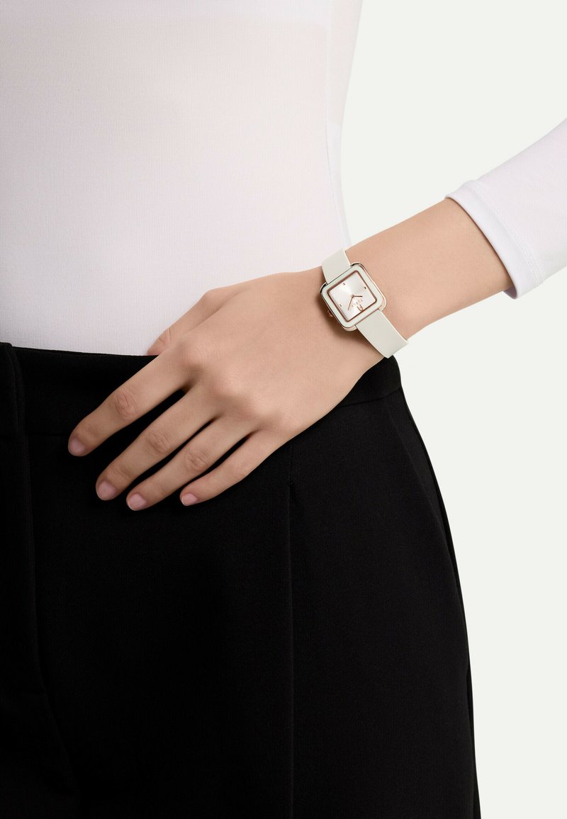 Montre carrée avec un bracelet en silicone blanc, des accents en or rose et un cadran blanc minimaliste. Portée sur une main reposant sur un pantalon noir.