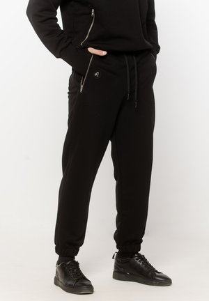 Osoba ubrana w czarne joggery z elastycznymi mankietami, czarne spodnie z kieszeniami na zamek błyskawiczny oraz czarne trampki, z rękami częściowo w kieszeniach, stojąca na białej podłodze.