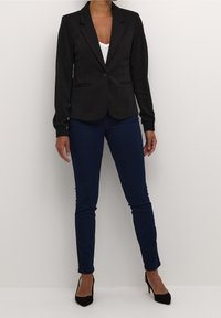 Czarna marynarka z jednym guzikiem, klapami i kieszeniami, noszona z niebieskimi spodniami typu slim fit i czarnymi szpilkami. Gładki materiał, dopasowany krój.