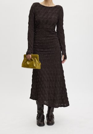 Donna che indossa un lungo abito nero testurizzato, stivali neri e tiene una piccola borsa verde oliva su uno sfondo chiaro e semplice.
