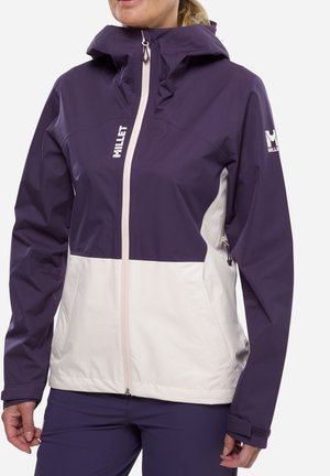 Femme portant une veste Millet violette et beige avec une fermeture éclair frontale et un logo sur la poitrine et la manche, debout devant un fond uni.
