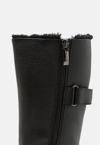 Botte en cuir noir avec une surface texturée, une bordure en fourrure en haut, une fermeture éclair sur le côté et un accent en sangle près de la cheville.