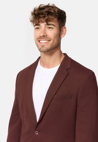 Brauner Blazer mit Reverskragen, Ein-Knopf-Verschluss und Taschenakzenten. Getragen über einem weißen T-Shirt, das eine glatte Textur aufweist.