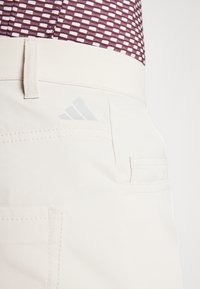 Vita shorts med en nära vy av en ficka, med subtil sömnad och en ljusgrå Adidas-logotyp på baksidan. Slät tygstruktur.