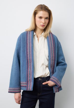 Giovane donna con capelli biondi indossa un cardigan blu testurizzato con ricami colorati sopra una camicia bianca e jeans scuri, in piedi contro un muro bianco.