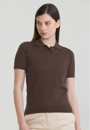 Ung kvinde med langt mørkt hår iført en brun polo med korte ærmer og beige bukser, ser mod venstre.