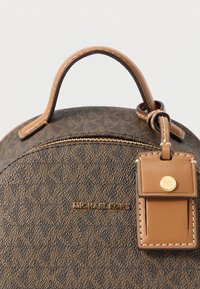 Brązowa torebka na przewieszenie z monogramem, zaokrąglonym kształtem, skórzanymi akcentami, złotymi okuciami i odpinaną metką, o fakturowanej powierzchni.