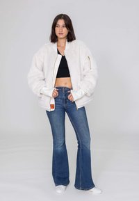 Witte, fuzzy bomberjack met een ritssluiting aan de voorkant, geribde manchetten en één zijzak. Draagt over een zwarte crop top en blauwe flared jeans.