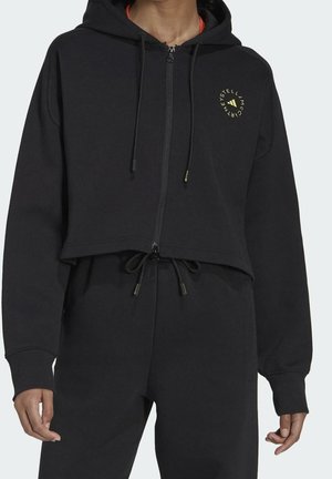 Persoon draagt een zwarte cropped zip-up hoodie met een cirkelvormig logo op de borst en bijpassende zwarte broek met trekkoord.