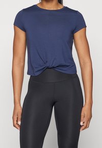 Camisa de manga corta de color azul marino con un detalle de nudo en la parte delantera, combinada con leggings negros de cintura alta. La tela parece suave y ligera.