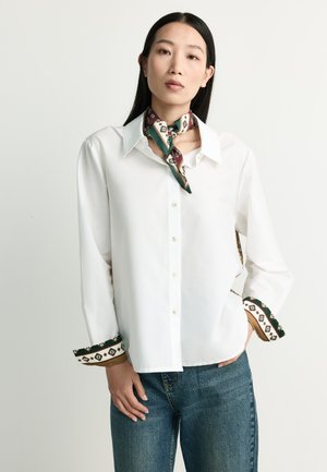 Woman with long black hair wearing a white buttoned shirt with patterned cuffs and collar detail, paired with blue jeans and a matching neck scarf.