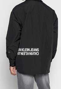 Black, lightweight, long-sleeve shirt with a relaxed fit. Features white text along the lower back, and button cuffs. Wrinkled texture.