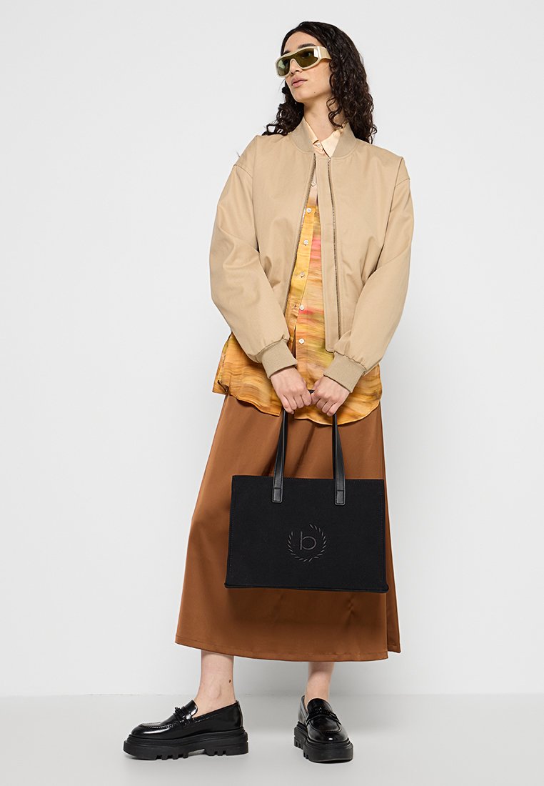 Beige Bomberjacke, bunte Musterbluse, braune Satin-Rock, schwarze Tragetasche mit Logo, klobige schwarze Schuhe, grüne Sonnenbrille.