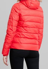 Rode pufferjas met capuchon, voorzien van een bontkraag. Het ontwerp heeft horizontale stiksels en elastische boorden. Draagt zwarte broek.