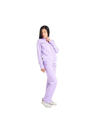Une femme porte un sweat-shirt mauve à fermeture éclair et un pantalon de jogging assorti, avec des baskets blanches. Elle regarde vers le bas en ajustant le col.