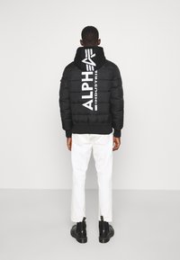 Schwarze Daunenjacke mit großem weißen "ALPHA INDUSTRIE"-Logo auf dem Rücken, eng anliegenden Bündchen und Kapuze. Weiße Hose und schwarze Stiefel runden den Look ab.