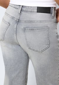 Gros plan sur une personne portant un jean gris clair avec des poches arrière et une étiquette de marque noire sur la ceinture, un haut blanc visible à la taille.