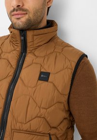 Gilet imbottito marrone con motivo esagonale, chiusura con zip e dettagli neri. Presenta un logo del marchio sul petto e uno strato interno scuro.
